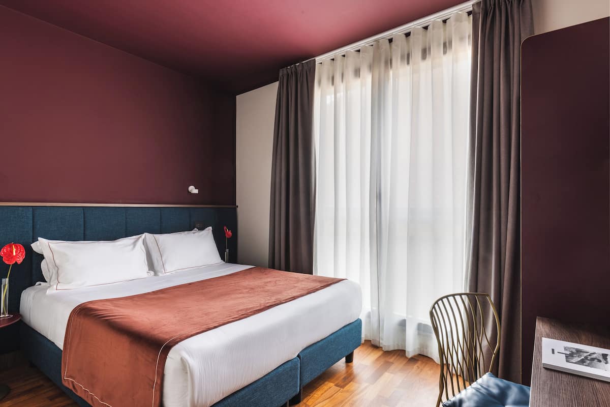 A neatly made bed with a brown blanket, a wooden nightstand with a vase of red flowers, and a chair by a window with curtains.