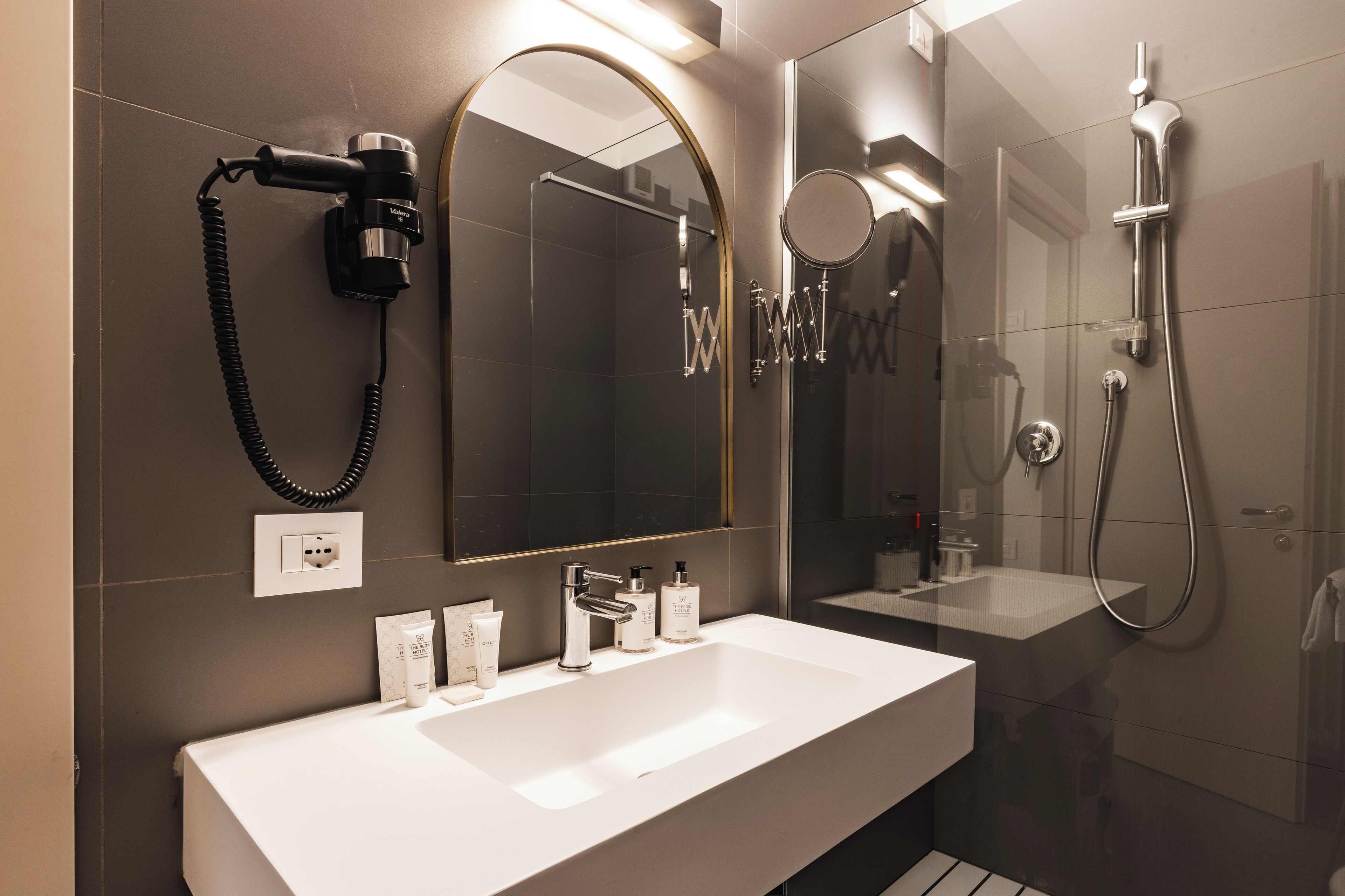 A modern bathroom with a large mirror, a white sink, a handheld showerhead, and a hairdryer.