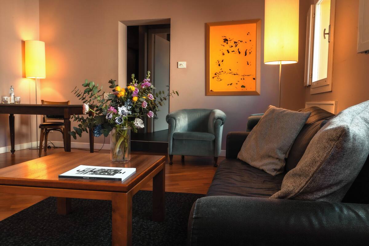 A living room with a wooden coffee table, a sofa, a chair, a vase with flowers, a lamp, and a painting on the wall.