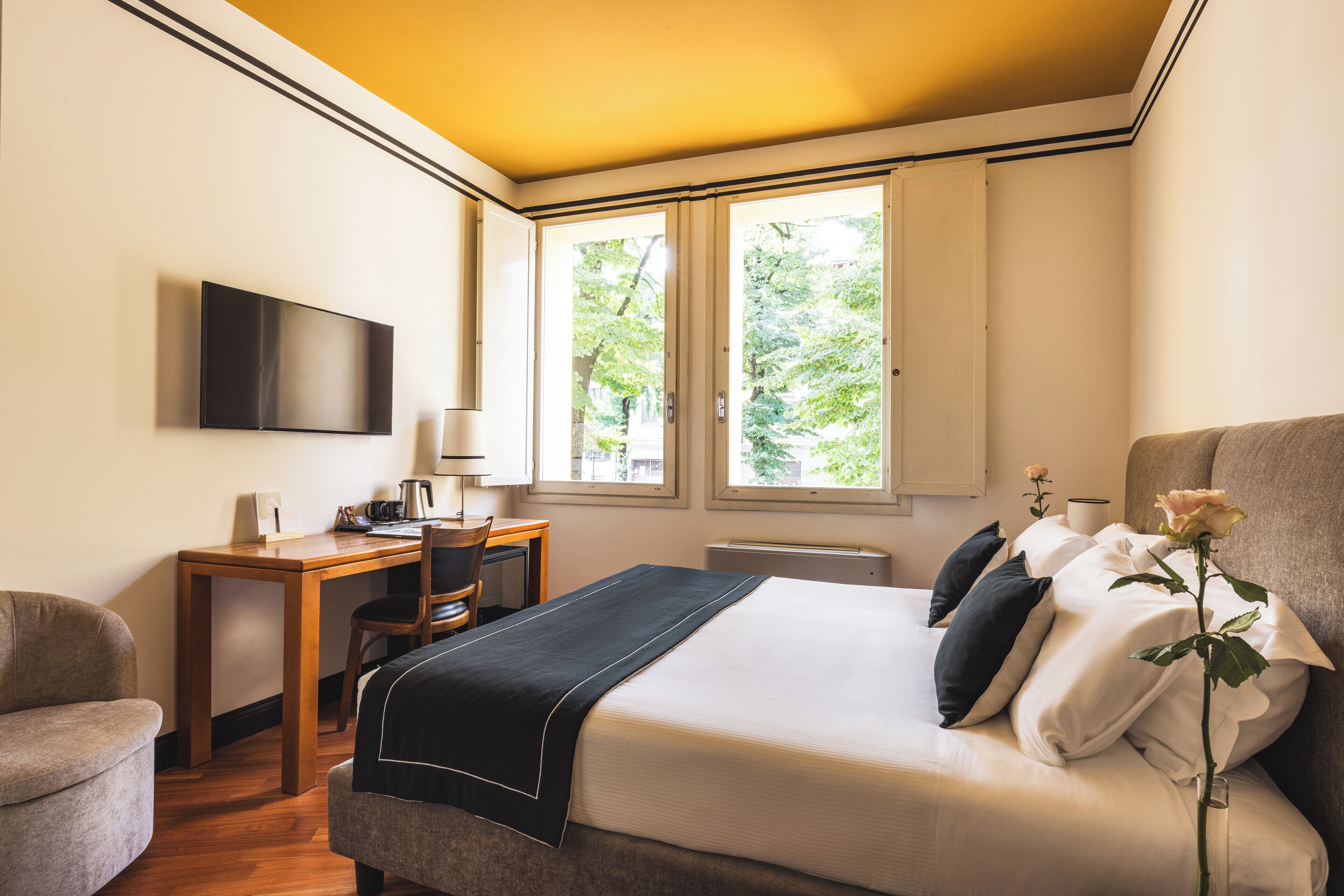 A hotel room with a bed, a desk, a chair, a television, and a window with a view of trees.