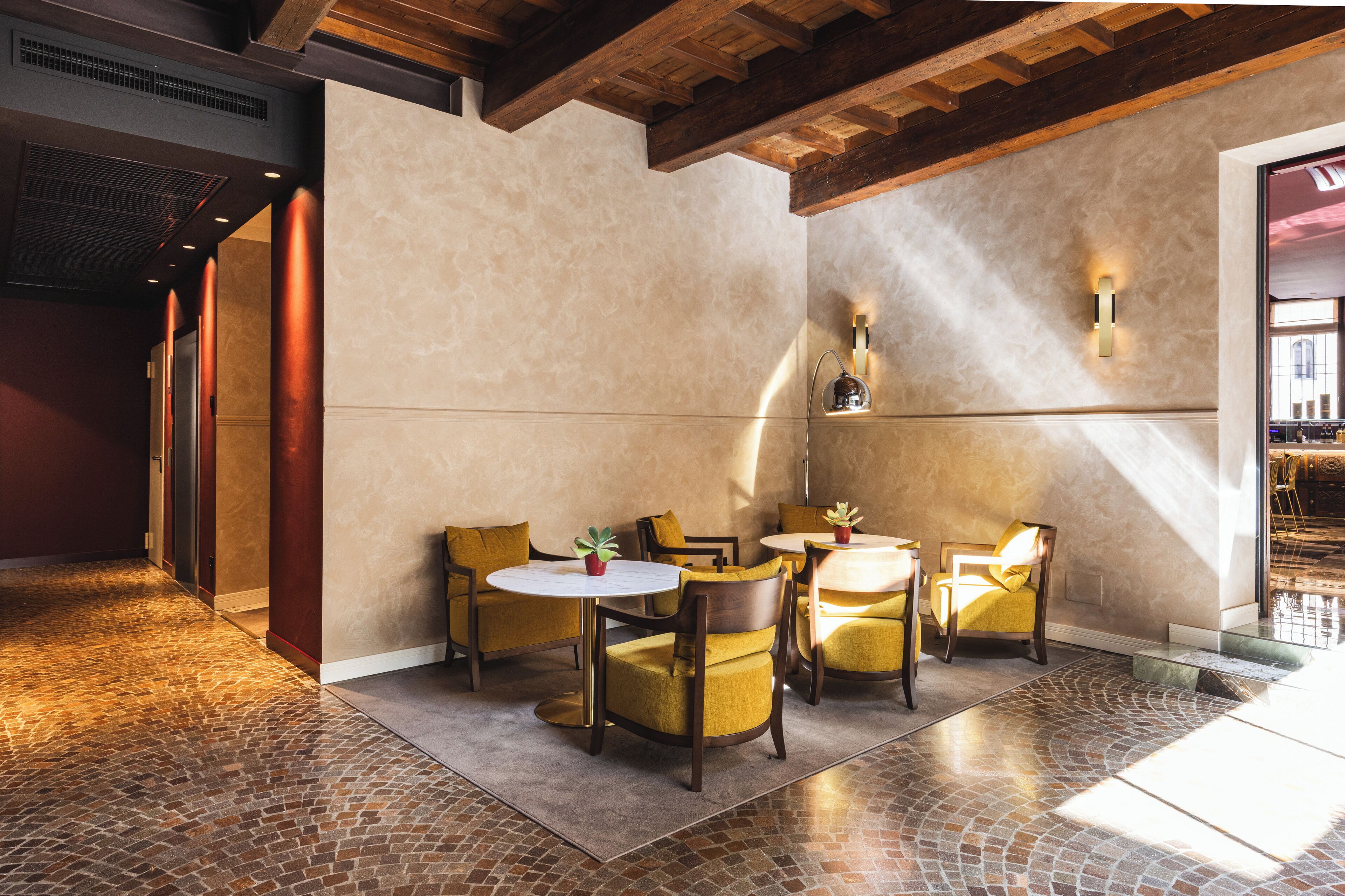 A modern interior with a brick floor, wooden ceiling beams, and a seating area with yellow chairs and a round table.