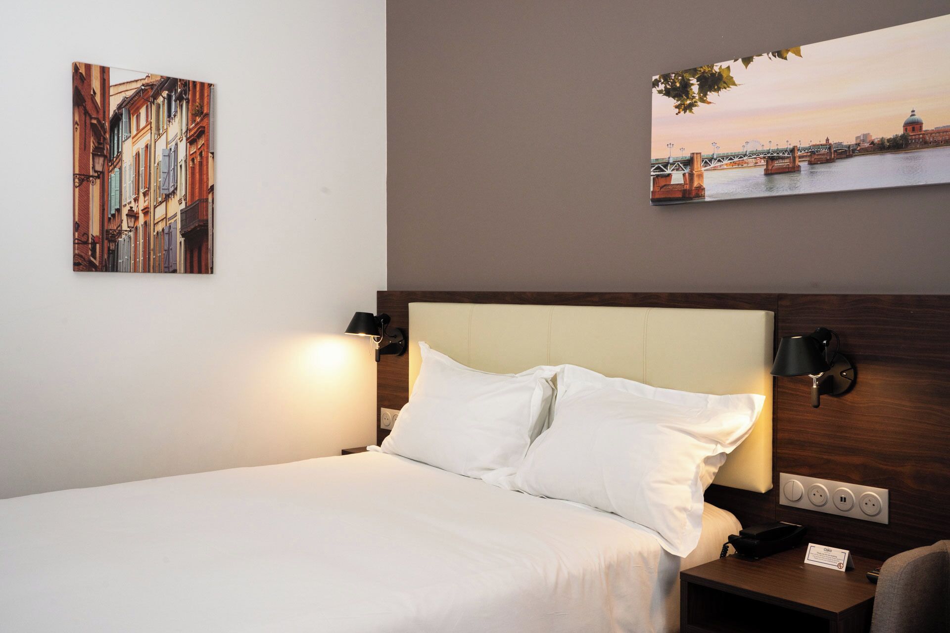 Une chambre d’hôtel comprenant un lit, deux lampes de chevet, une table de chevet avec un téléphone et deux œuvres d’art fixées au mur représentant des scènes urbaines.