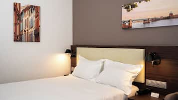 Une chambre d’hôtel comprenant un lit, deux lampes de chevet, une table de chevet avec un téléphone et deux œuvres d’art fixées au mur représentant des scènes urbaines.