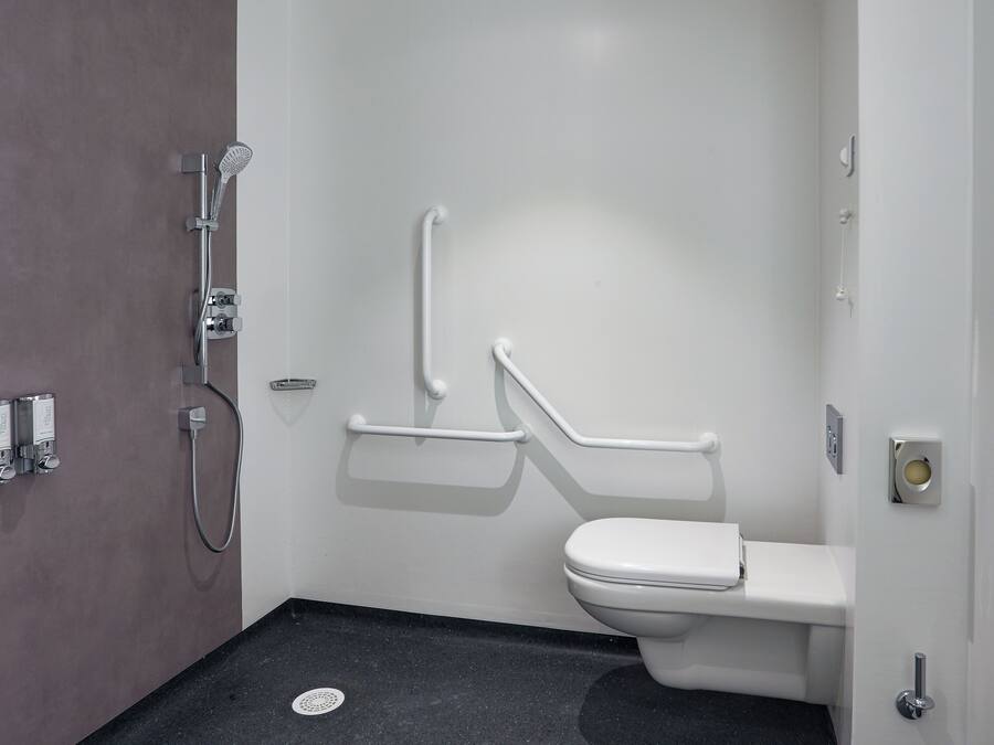 Un baño con inodoro blanco, ducha con cabezal de ducha manual y barra de apoyo.