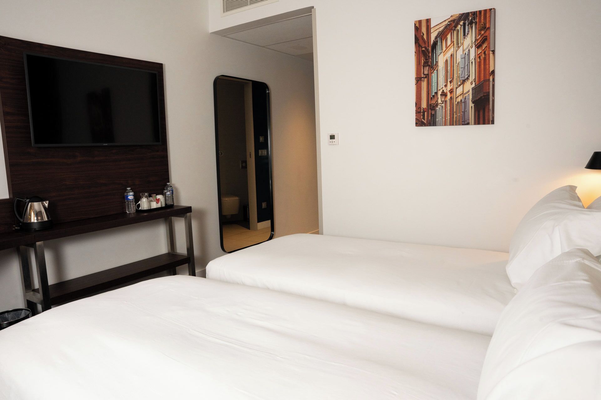 A hotel room with a bed, a TV, a mirror, and a painting on the wall.