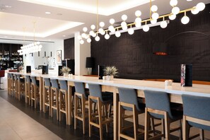A modern bar area with a long counter, multiple bar stools, and a decorative light fixture. - OSKO - Hôtel Toulouse Aéroport (Blagnac)