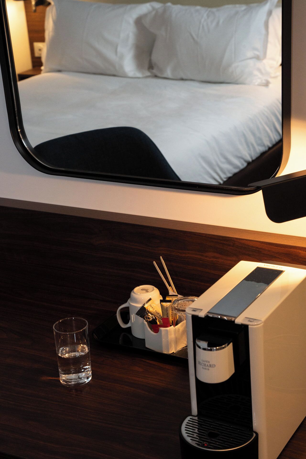 Une chambre d’hôtel comprenant un lit, une table de chevet avec un verre d’eau, une machine à café et un porte-tasse avec des ustensiles.