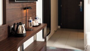 Habitación de hotel con televisor de pantalla plana montado en un panel de madera, cafetera, hervidor de agua y dispensador de agua sobre una consola de madera. Hay una puerta con un cartel y una alfombra estampada en el suelo.