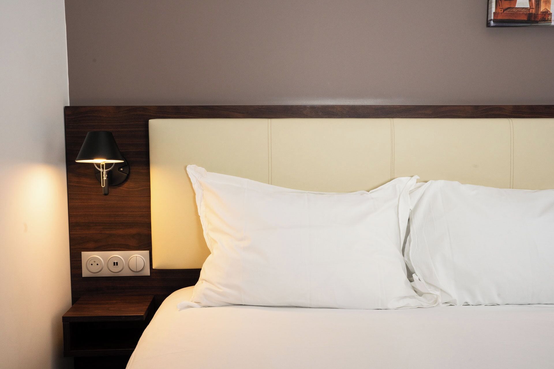 Una cama bien hecha con sábanas blancas, un cabecero de madera y una lámpara fijada a la pared.