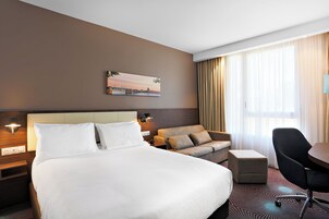 A hotel room with a large bed, a sofa, a desk, and a chair. There is a painting on the wall and a window with curtains.