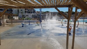 Un espace agrémenté d’une fontaine avec de nombreux jets d’eau, une pergola en bois au-dessus et des bâtiments en arrière-plan.