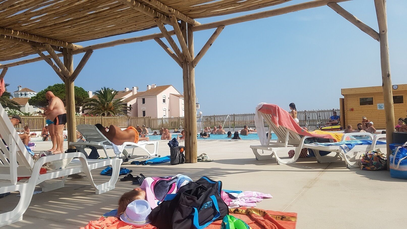 Ein Poolbereich am Strand mit Liegestühlen und Sonnenschirmen, umgeben von Gebäuden und einem klaren Himmel.