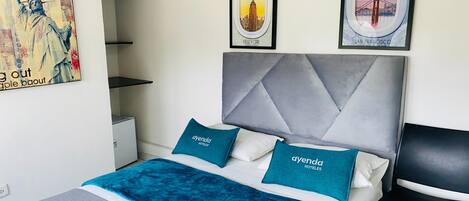 Habitación de hotel con una cama, cabecera, dos almohadas con la marca 'ayenda hotels' y dos cuadros con imágenes de lugares emblemáticos de Nueva York.