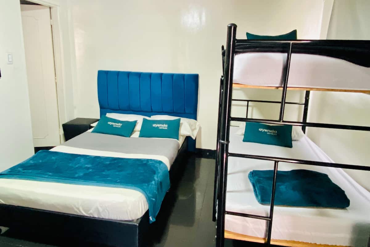 Una habitación con una cama individual con cabecera y almohadas azules a juego, una cama litera con una tonalidad similar, una mesita de noche y una puerta.