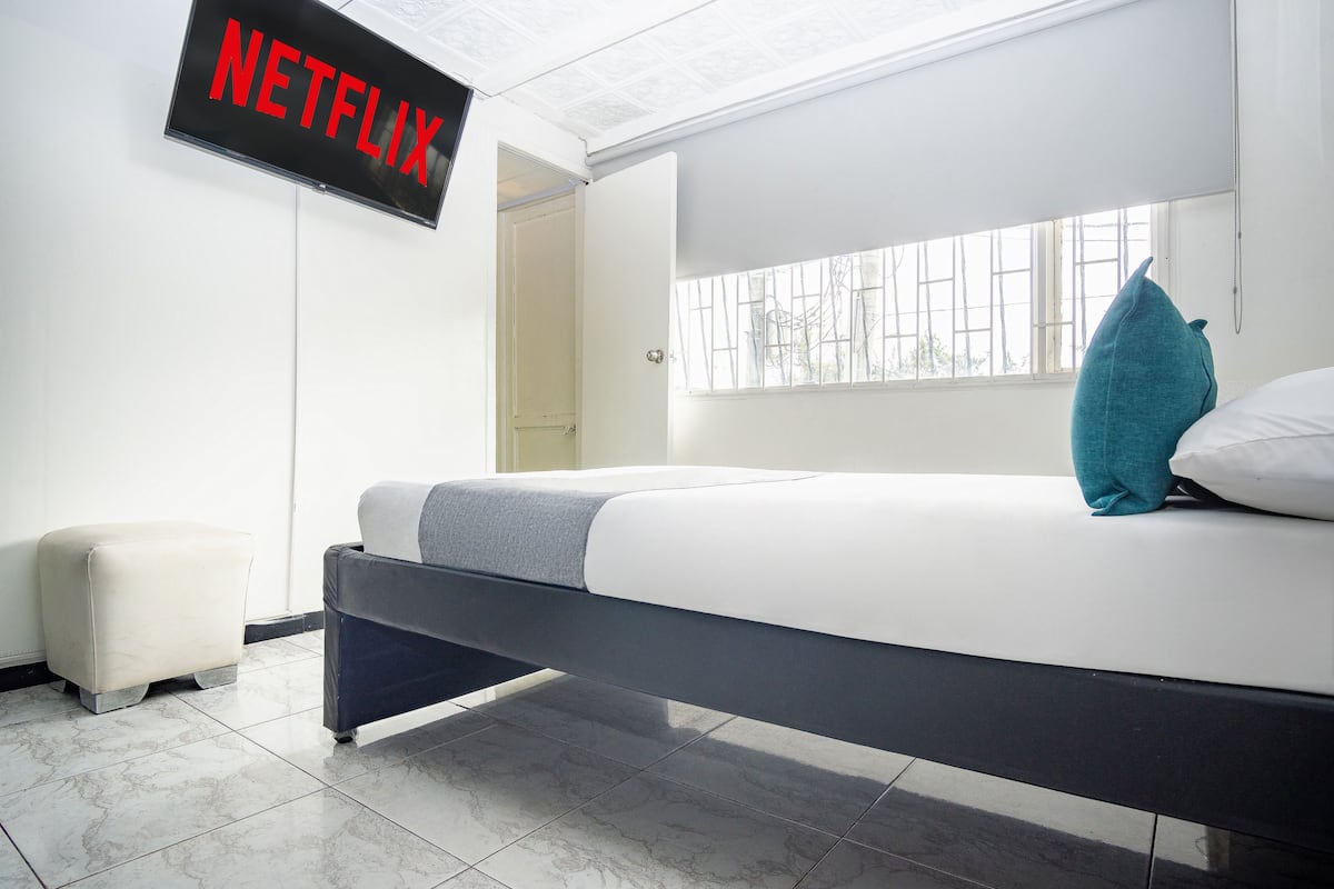 Un dormitorio moderno con una cama, una mesita de noche pequeña, un televisor de pared que muestra 'NETFLIX' y una ventana con vistas al exterior.
