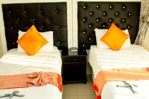 A hotel room with two beds, each with a black tufted headboard and white bedding. There are two orange pillows on each bed.