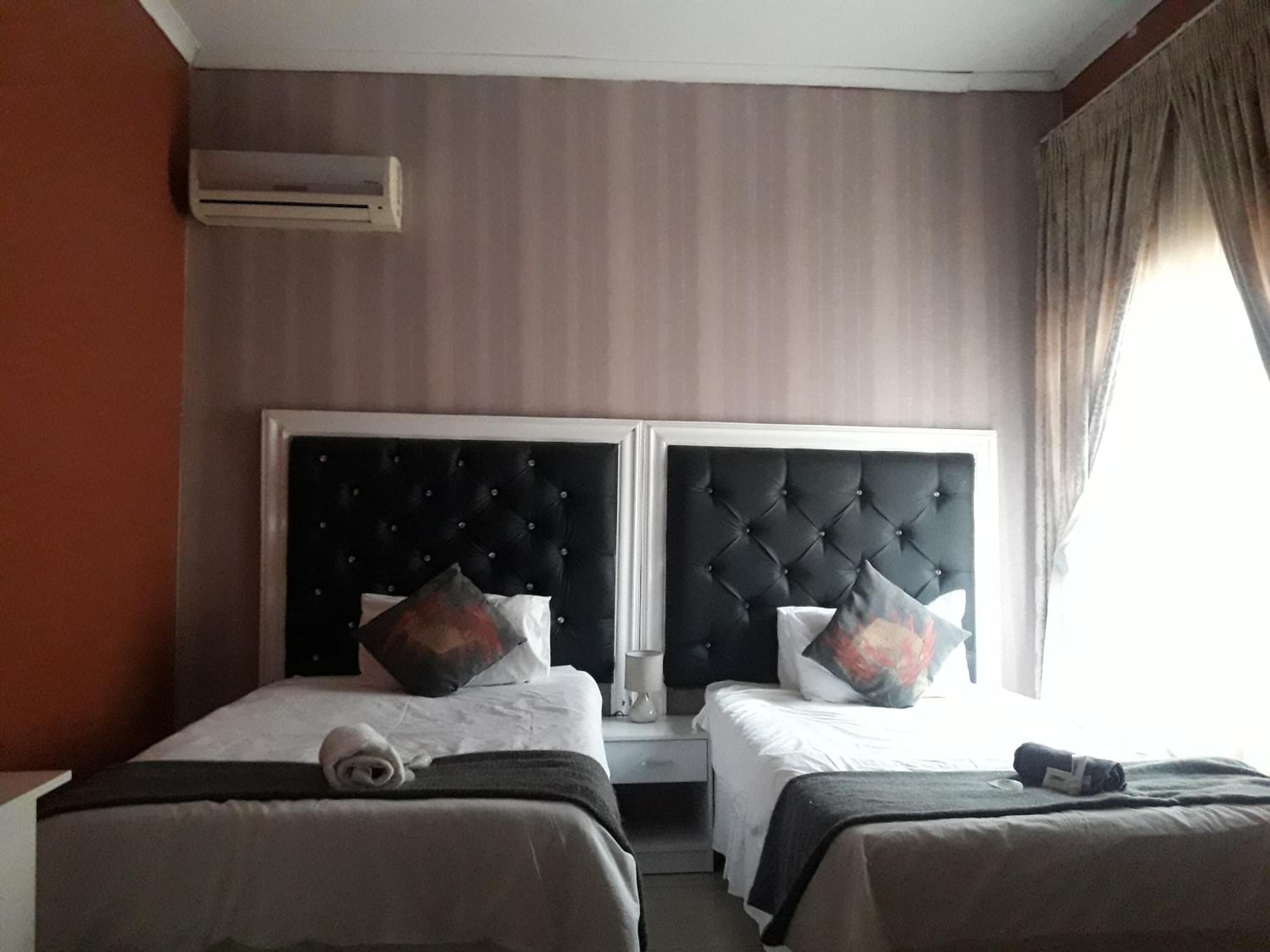 A hotel room with two beds, each with a black tufted headboard, white bedding, and decorative pillows. An air conditioning unit is mounted on the wall, and a window with curtains is visible.