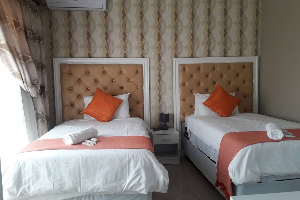 Een hotelkamer met twee bedden, elk voorzien van een gestuikt hoofdeinde, witte beddengoed en oranje kussens. Elk bed heeft een nachtkastje met een lamp en een opgevouwen handdoek.