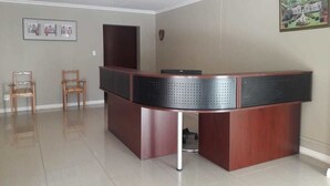 A reception desk with a dark wooden finish, two wooden chairs, a framed picture on the wall, and a tiled floor.
