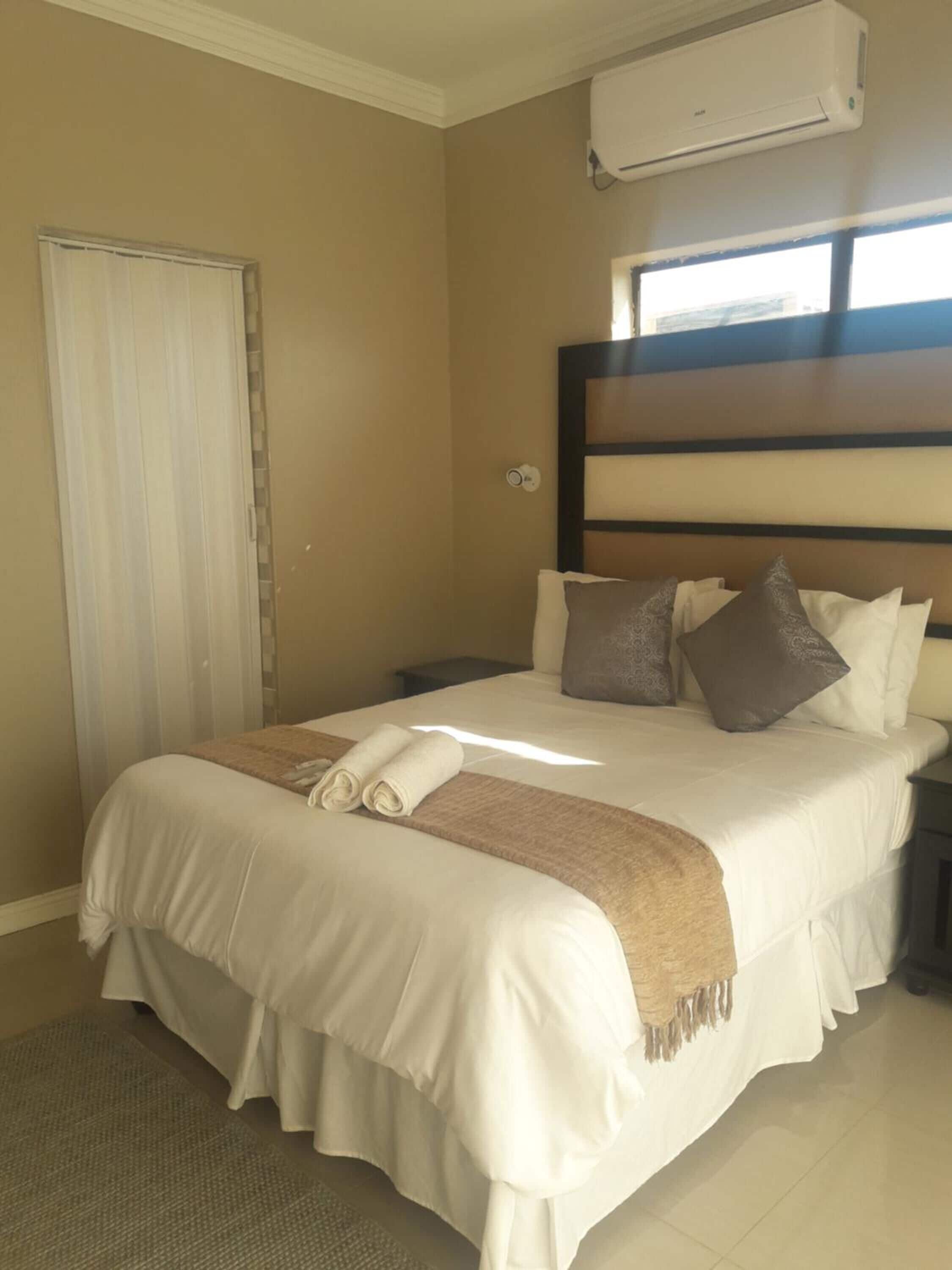 Cama bem arrumada com um edredom branco e castanho, duas almofadas e uma toalha enrolada. Há uma mesinha de cabeceira com abajur, uma janela com persianas e um ar condicionado na parede.