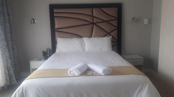 A hotel room with a large bed, white bedding, a headboard with a geometric design, two bedside tables, and a curtain on the left.
