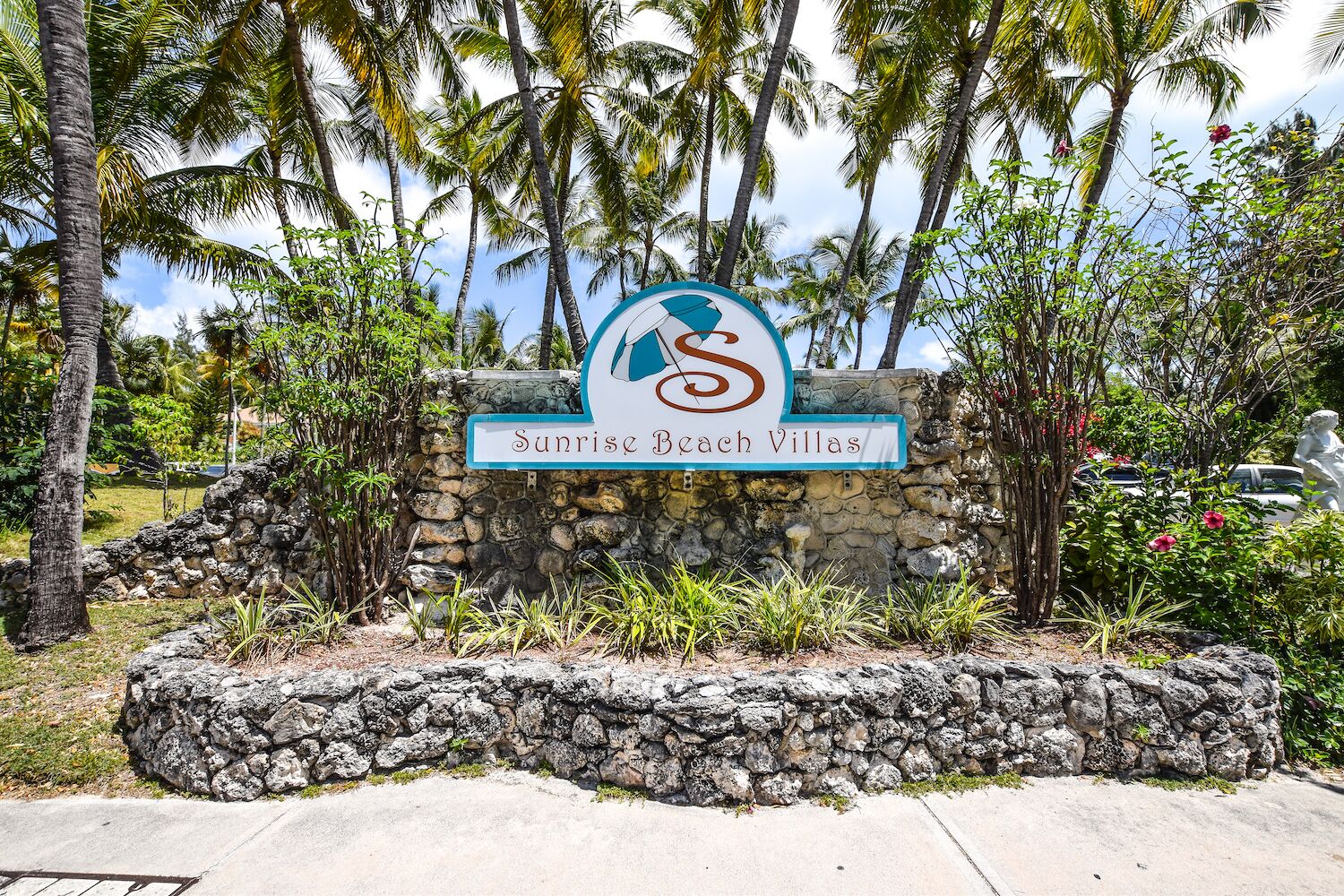 A sign for Sunrise Beach Villas with a logo, surrounded by palm trees and a stone wall.
