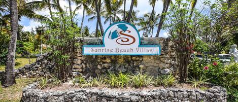 A sign for Sunrise Beach Villas with a logo, surrounded by palm trees and a stone wall.