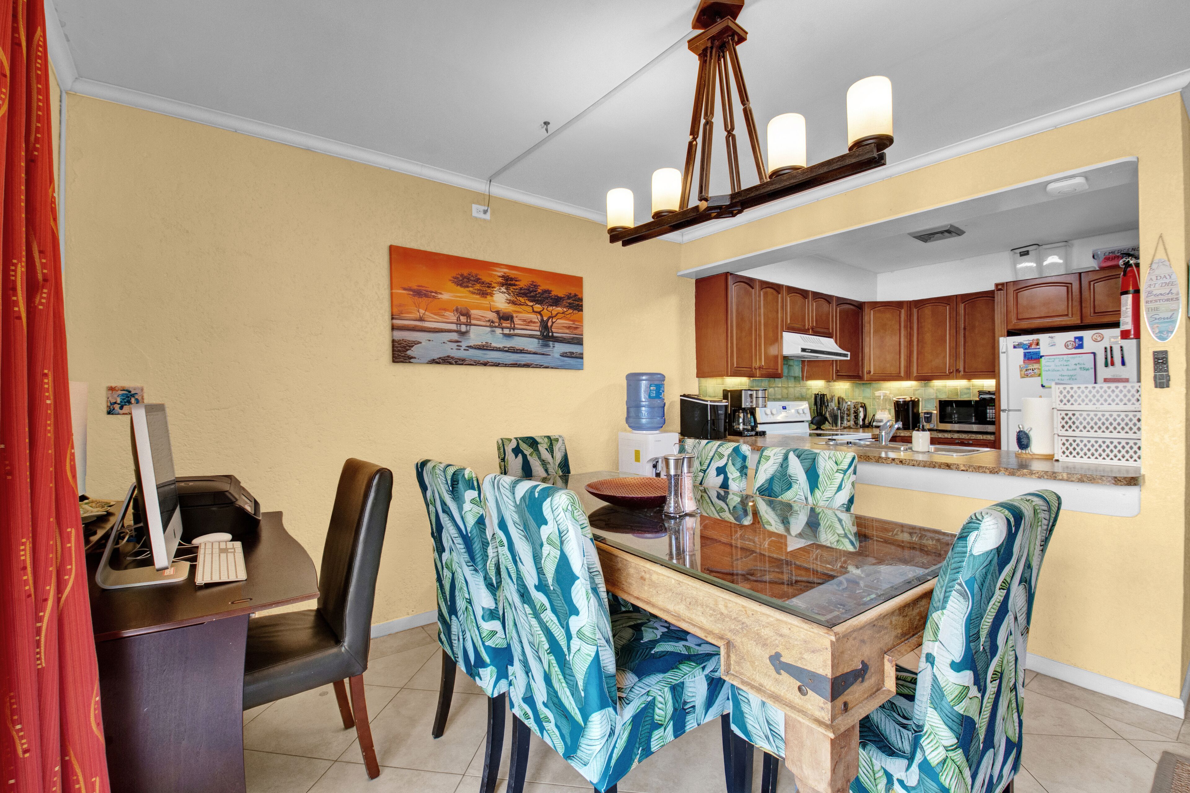 A dining area with a wooden table, chairs with tropical leaf patterns, a painting of a sunset, and a kitchen in the background.