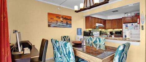 A dining area with a wooden table, chairs with tropical leaf patterns, a painting of a sunset, and a kitchen in the background.