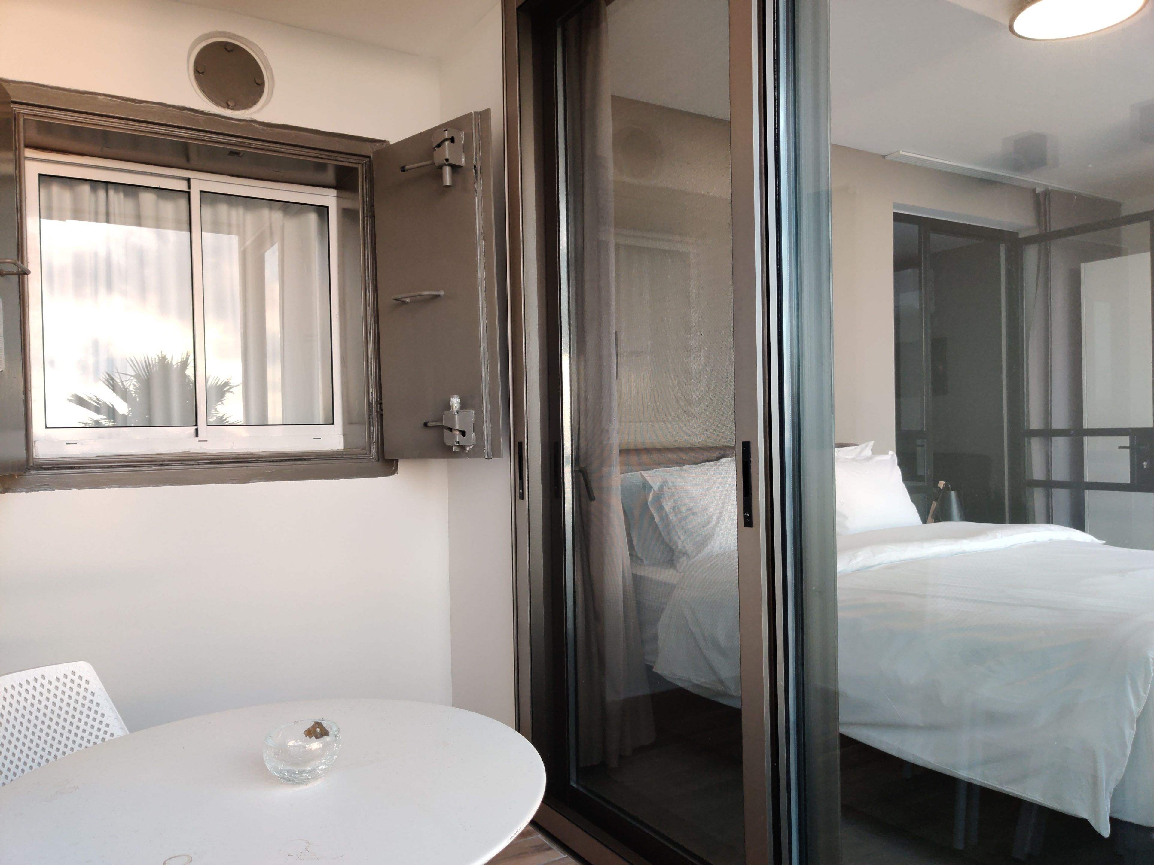 A hotel room with a sliding glass door leading to a balcony, a bed with white linens, a small round table with a glass object, and a window with curtains.