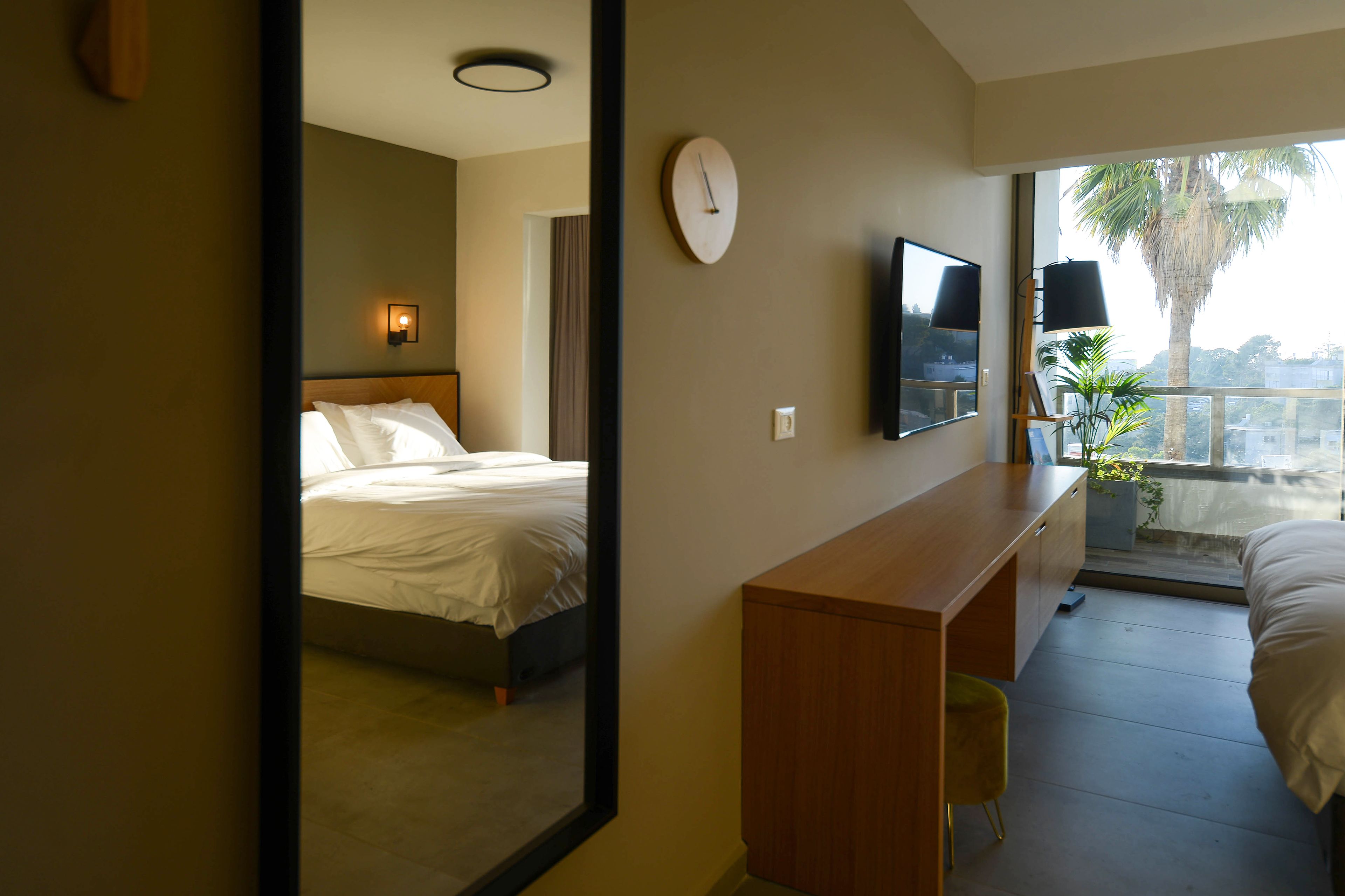 A hotel room with a bed, a desk, a TV, a clock, and a balcony with a plant.