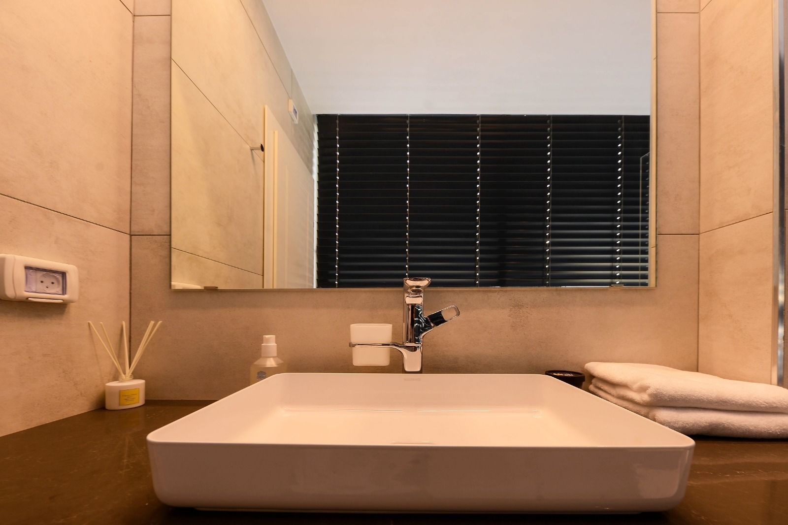 A modern bathroom with a large rectangular sink, a mirror above it, and a window with blinds.
