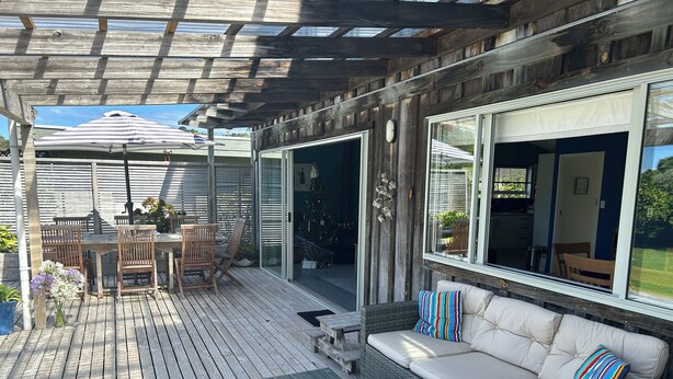A covered outdoor patio with a wooden deck, a dining table with chairs, a sofa with cushions, and a large umbrella. - Coro Beach Haven - Hahei Holiday Home (Hahei)