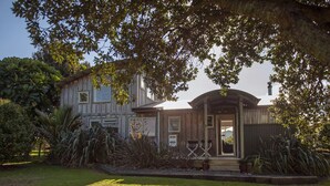 A two-story house with a covered porch, surrounded by a well-maintained lawn and various plants. - Coro Beach Haven - Hahei Holiday Home (Hahei)