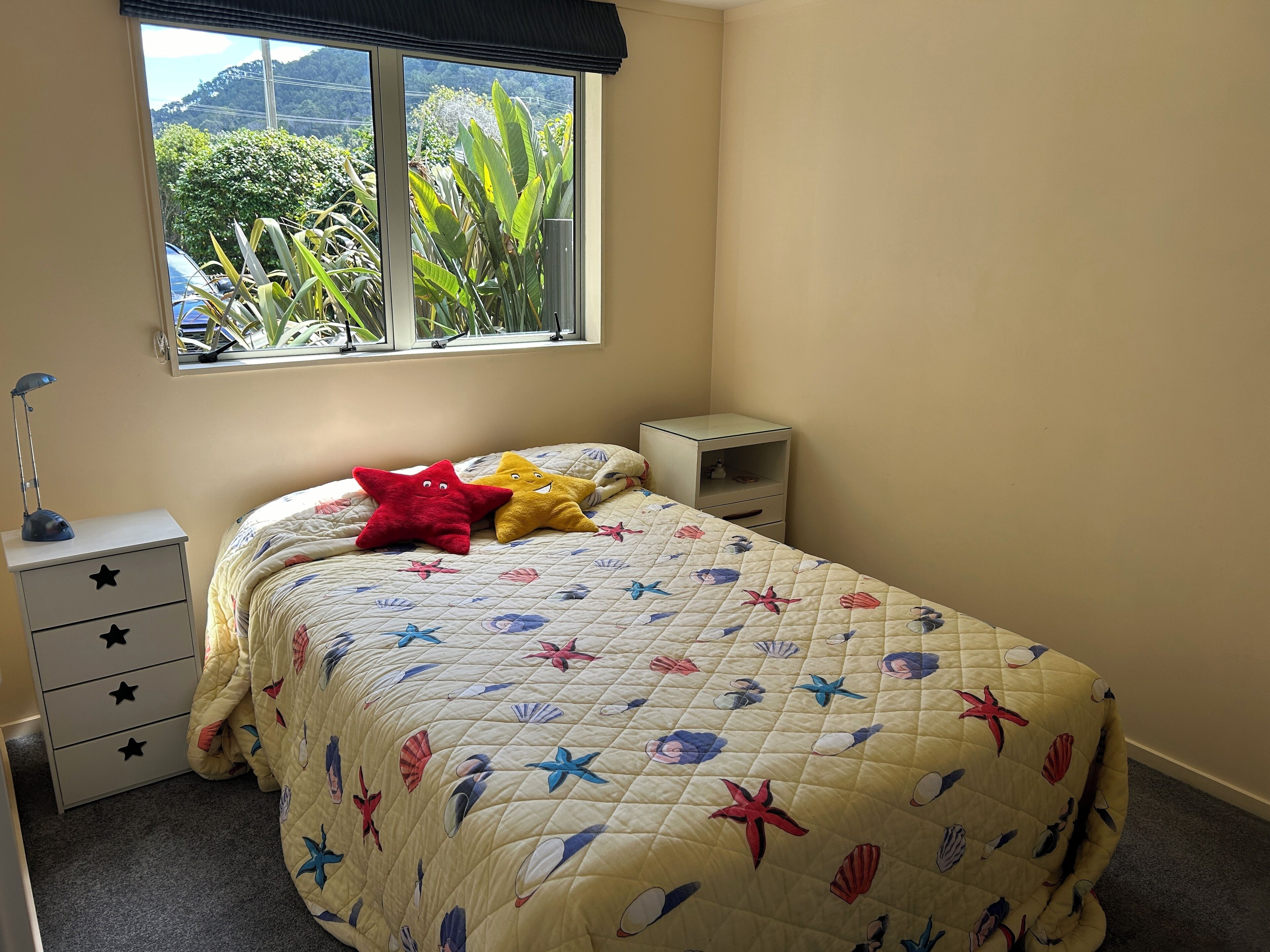 A bedroom with a bed, a nightstand, a bedside table, a window, and a view of greenery outside.
