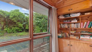 A wooden cabin with a large window, a bookshelf with various books and games, and a view of greenery outside. - Little Lockwood - Kuratau Bach (Kuratau)