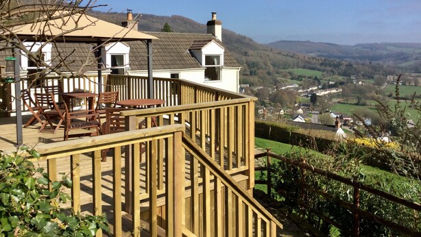 Une terrasse en bois, agréée de mobilier d’extérieur, qui offre une vue imprenable sur une vallée pittoresque, avec ses maisons et ses collines.