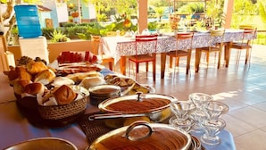 Una mesa de buffet con diversos alimentos y recipientes, ubicada en un comedor al aire libre con vistas a la vegetación.