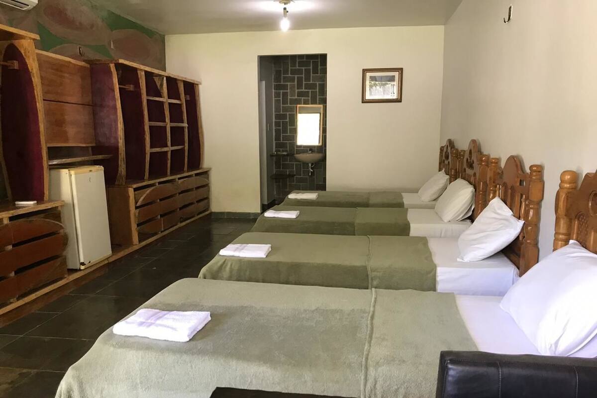Habitación de hotel con cuatro camas dispuestas en fila, un lavamanos pequeño y un armario de madera.