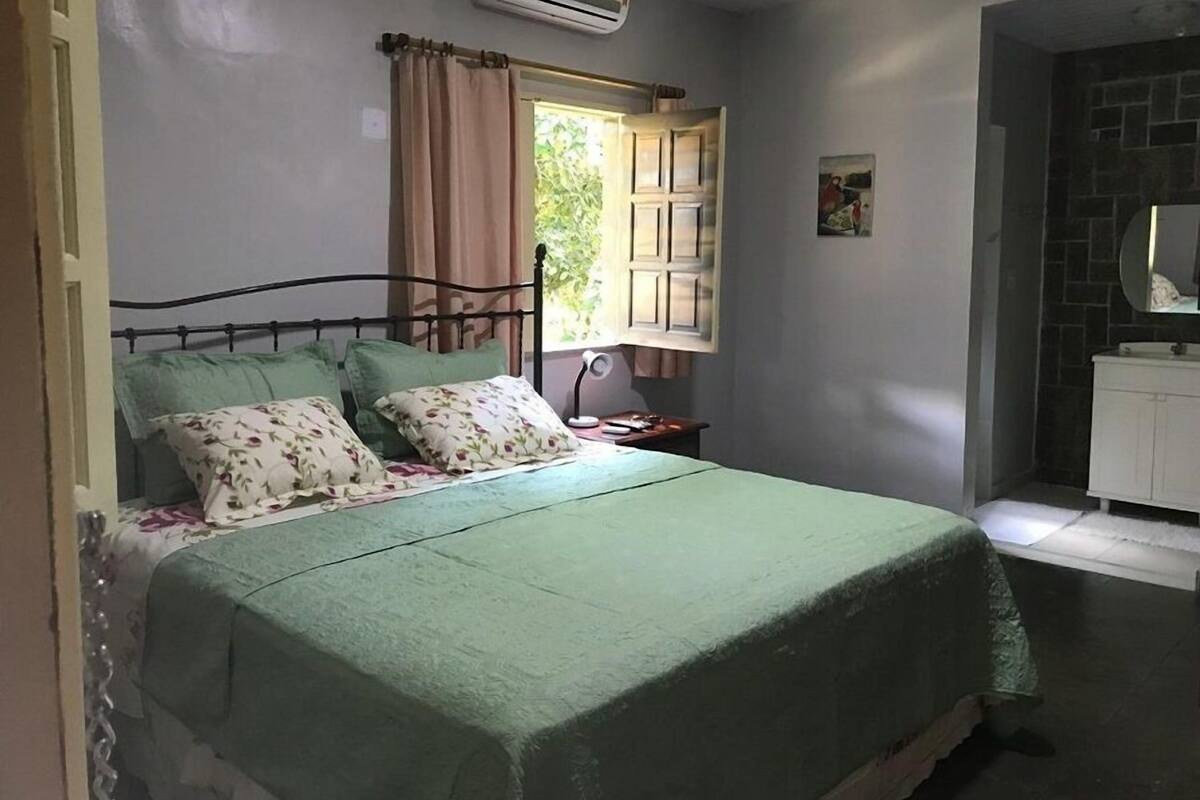 Un dormitorio con cama, mesita de noche, equipo de aire acondicionado, ventana con cortinas y baño con lavabo y espejo.