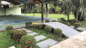 Una cancha deportiva al aire libre con zona de estar cubierta, rodeada de follaje y palmeras.