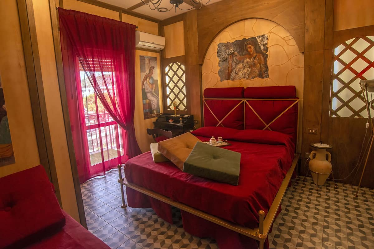 Une chambre avec un lit rouge, une petite table avec une lampe et une fenêtre donnant sur un beau paysage.