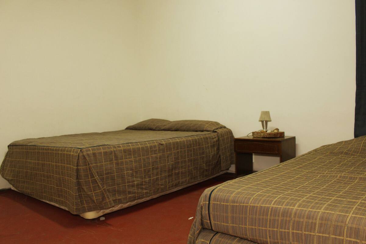 A room with two beds, each with a checkered bedspread, a nightstand with a lamp, and a plain wall.