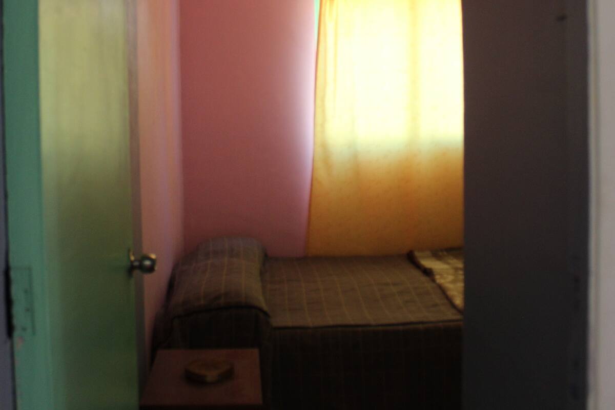 A small, dimly lit room with pink walls, a single bed, a bedside table, and a window with curtains.