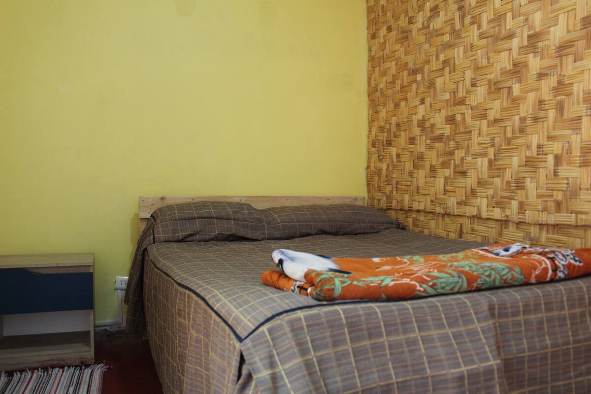 A bed with a checkered bedspread, a woven headboard, and a striped rug.
