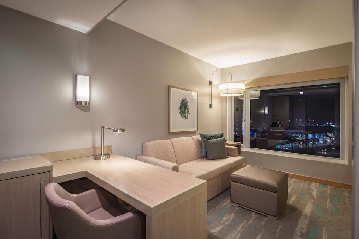 Una habitació d'hotel moderna amb un sofà, escriptori, cadira, finestra amb vista a la ciutat i una obra d'art emmarcada a la paret.