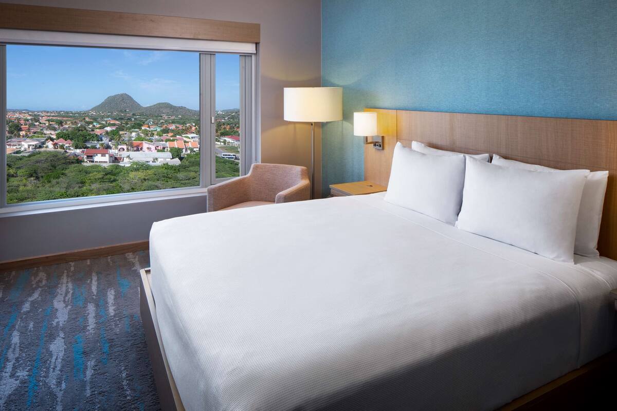 Habitació d'hotel amb un llit gran, una cadira, una tauleta de nit i vista a una vila i muntanyes.