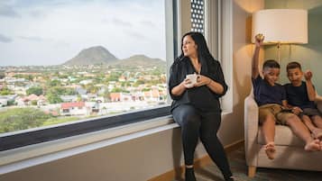 A woman sitting by a window with a view of a town and a mountain, two children on a couch, and a lamp on the wall.