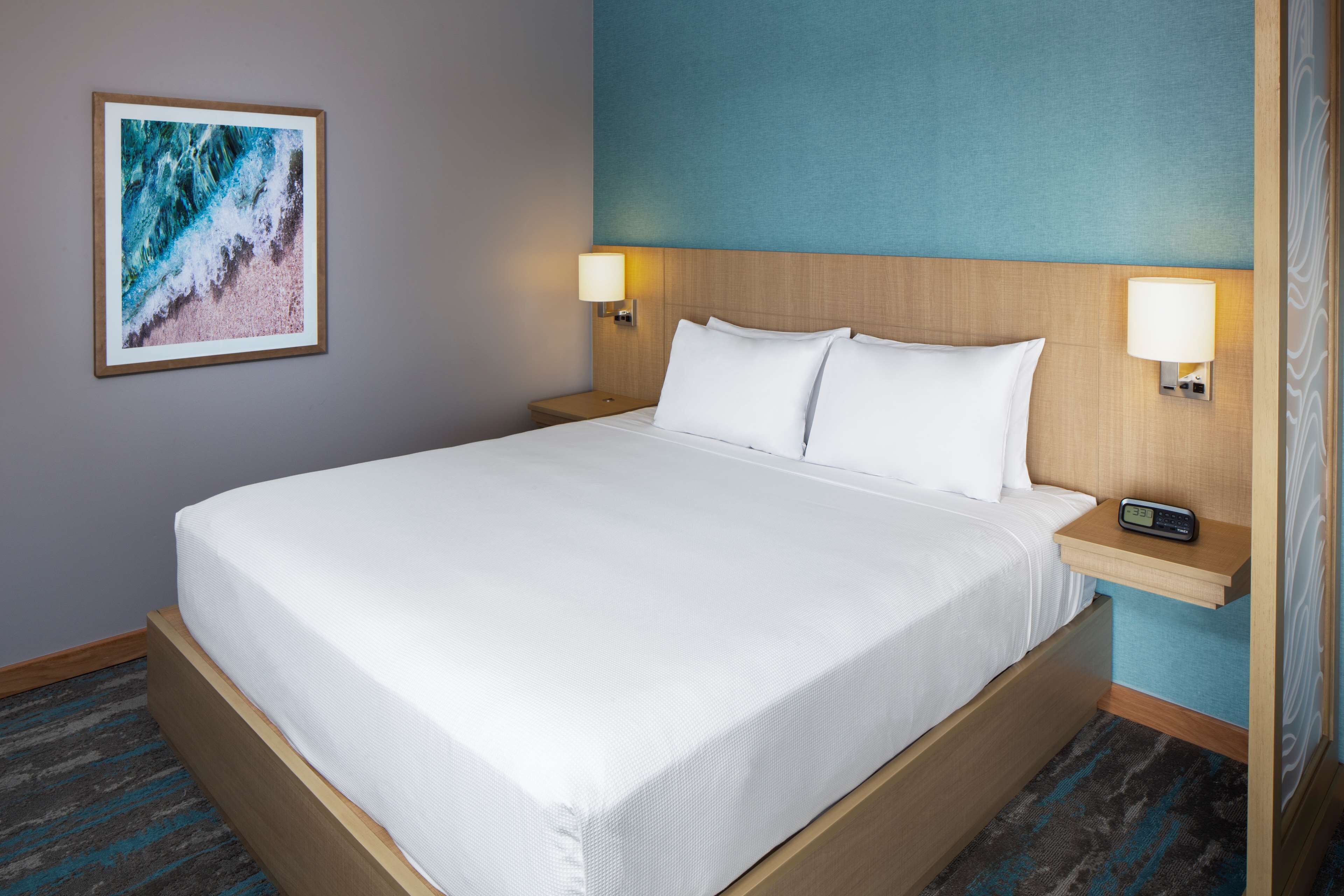 A neatly made bed with white linens, two bedside lamps, and a framed picture of a beach scene on the wall.