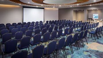Un salón de conferencias con filas de sillas azules, una pantalla grande y un diseño de techo de madera.